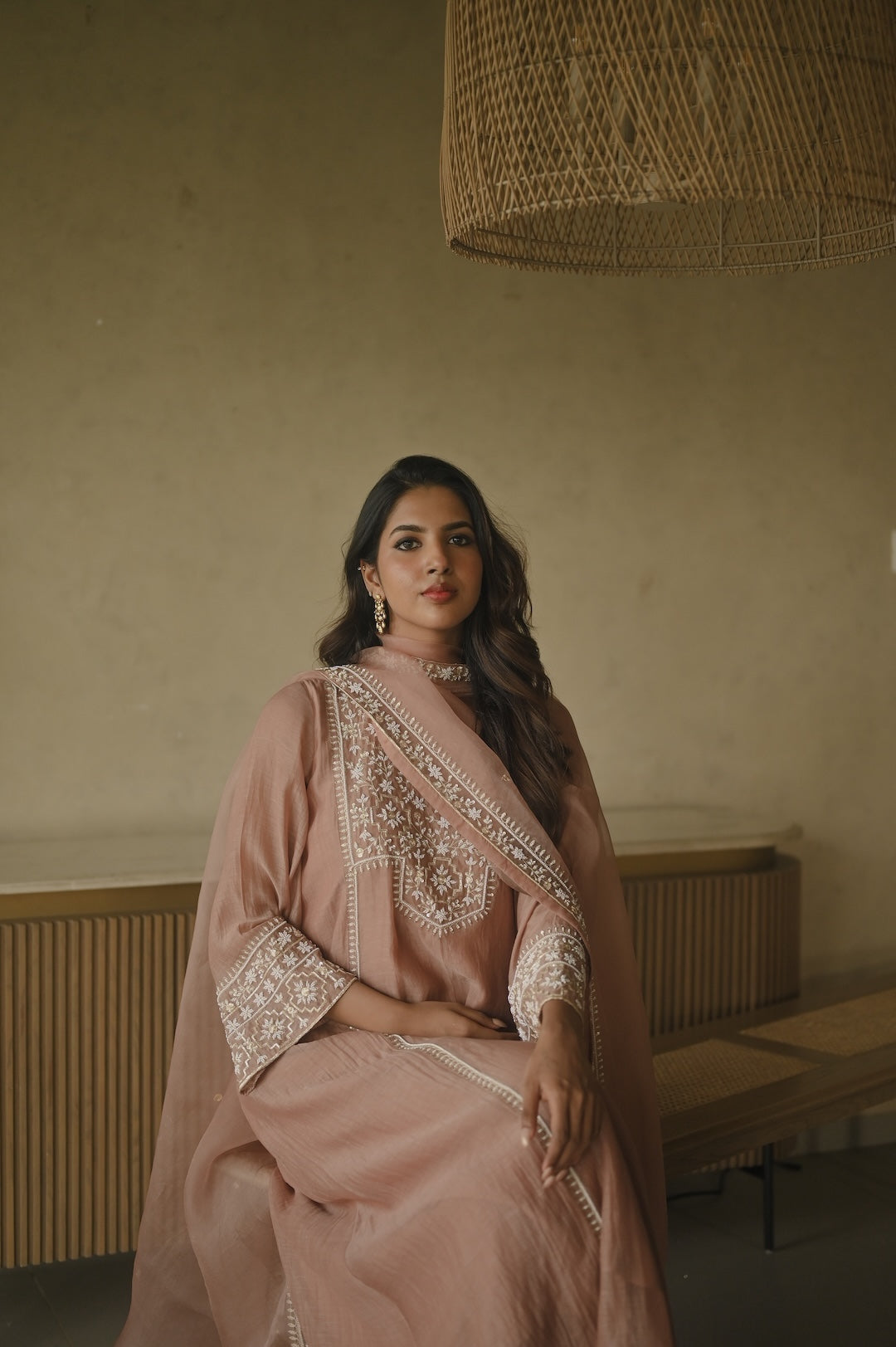 Woman in a pink traditional outfit with intricate patterns sitting in a room with a textured wall and wooden bench.